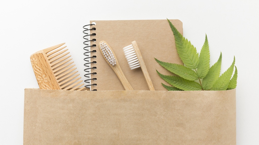 Exemplo de Kit Ecológico com sacola reciclável, caderneta com capa e folhas reciclados, escova de dente de madeira, pente de madeira e folha de planta.